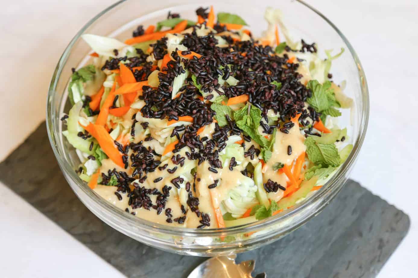 accompaniment salad with cabbage carrots ginger miso dressing and forbidden black rice and sesame seeds in a glass serving bowl with a serving spoon and fork