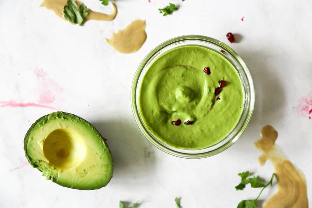 horizontal view of green tahini sauce in a bowl next to an avocado