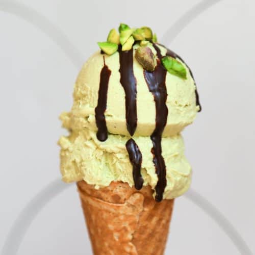 picture of vegan pistachio ice cream on a waffle cone with chopped pistachio nuts and chocolate syrup