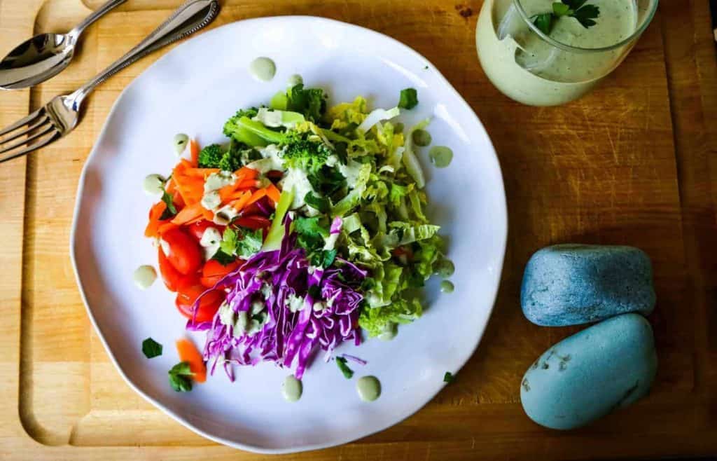 rainbow salad with vegan green goddess dressing on a white plate on a cutting board with carrots tomatoes purple cabbage broccoli romaine lettuce and herbs
