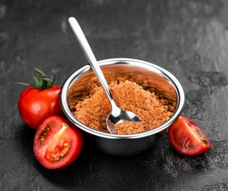 tomato powder in a metal bowl with a spoon surrounded by sliced tomatoes