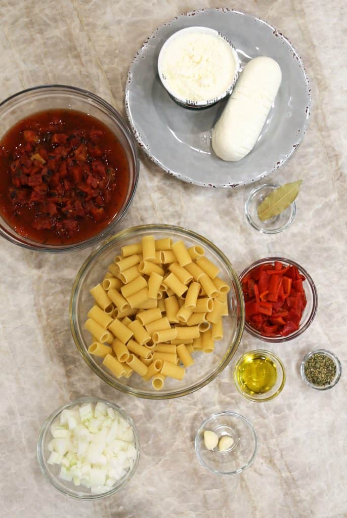 ingredients for gluten free stuffed rigatoni in clear bowls including onion, rigatoni, olive oil, Italian seasoning, red peppers, garlic cloves, roasted tomatoes, ricotta cheese and mozzarella cheese
