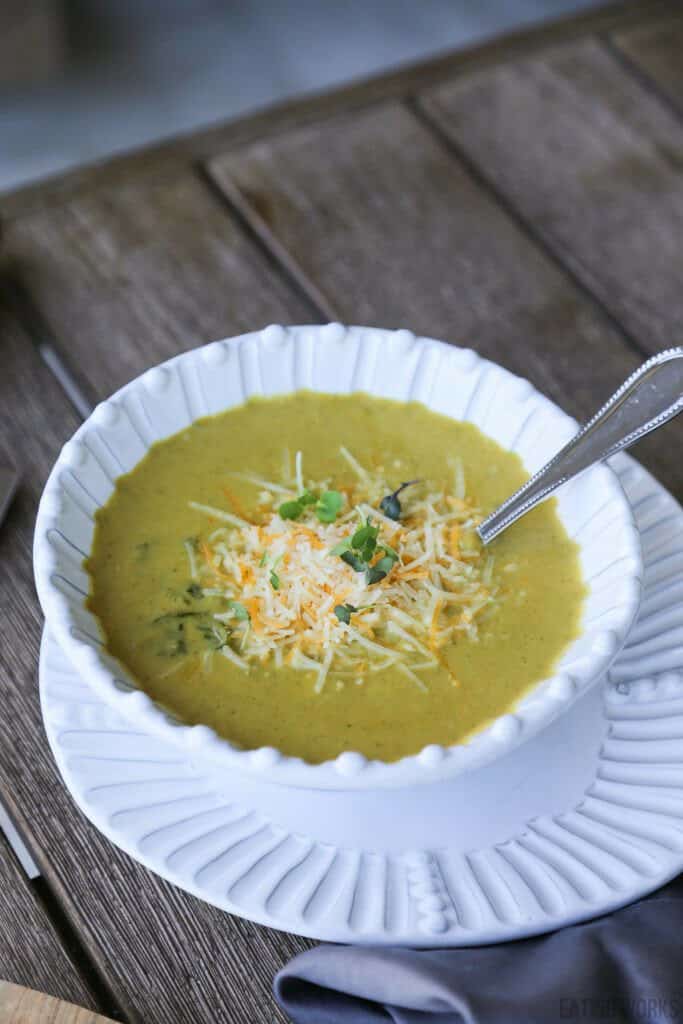 gluten free broccoli cheddar soup in a bowl garnished with cheese and greens on a table with a napkin pot of soup and a knife and two spoons 