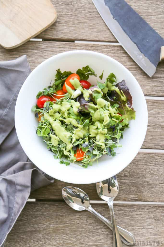 vegan avocado dressing recipe drizzled over a salad with micro greens and tomatoes in a white bowl with spoons and a napkin