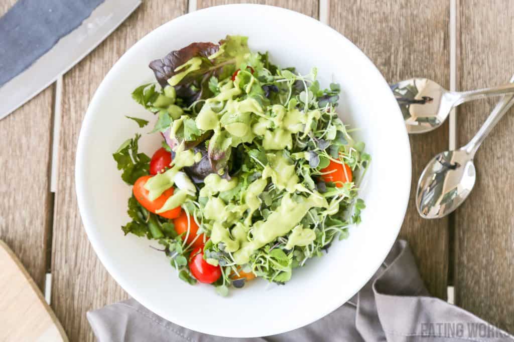 vegan avocado dressing recipe drizzled over a salad with micro greens and tomatoes in a white bowl with spoons and a napkin