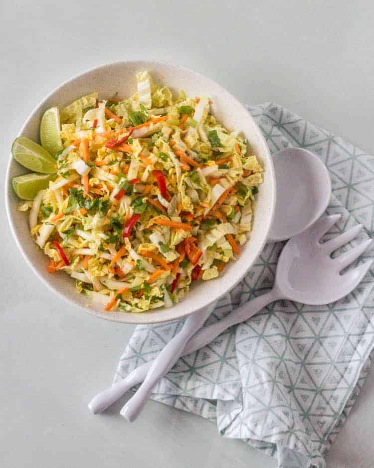 A round white ceramic bowl with Southwest Coleslaw on a light background with an aqua and white napkin and salad servers.