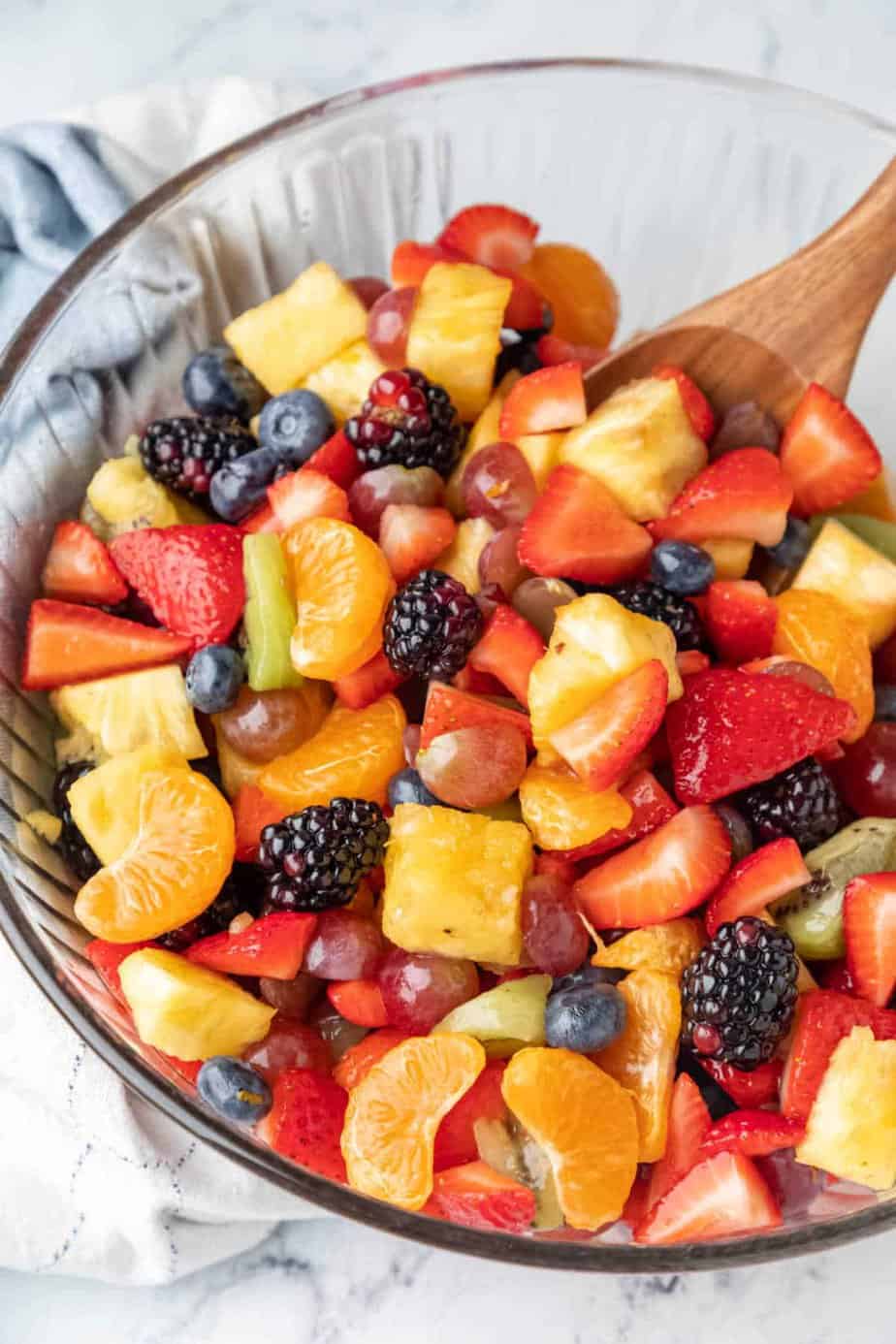 A wooden spoon in a glass mixing bowl full of fruit salad. 