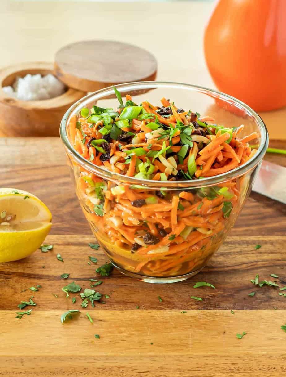 A clear glass bowl of Moroccan Carrot Salad with Lemon Dressing, a wood salt box and bottle of olive oil.