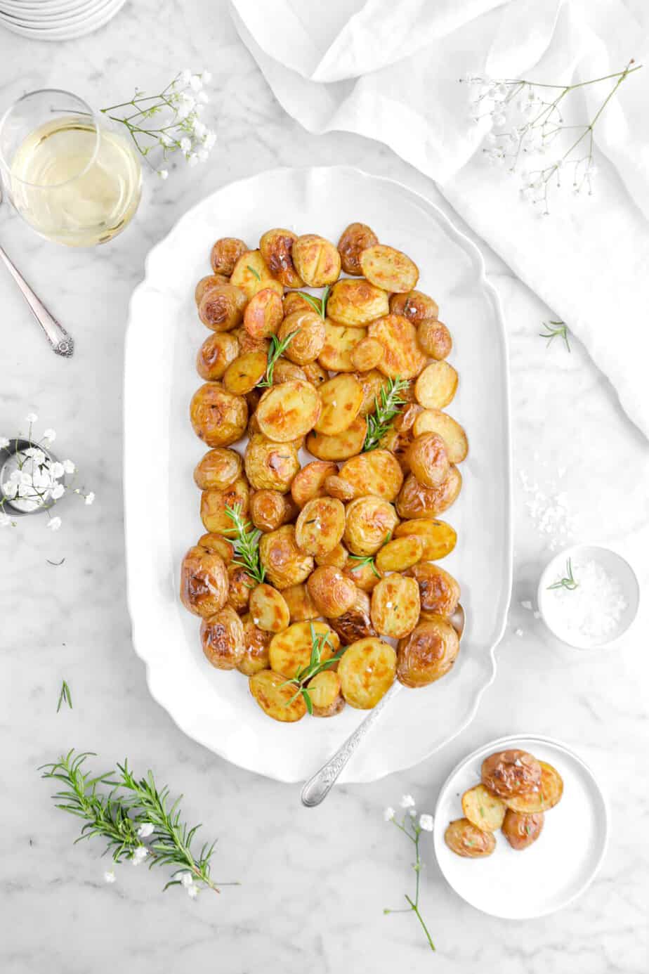 Roasted potatoes on white platter with spoon and fresh rosemary sprigs.