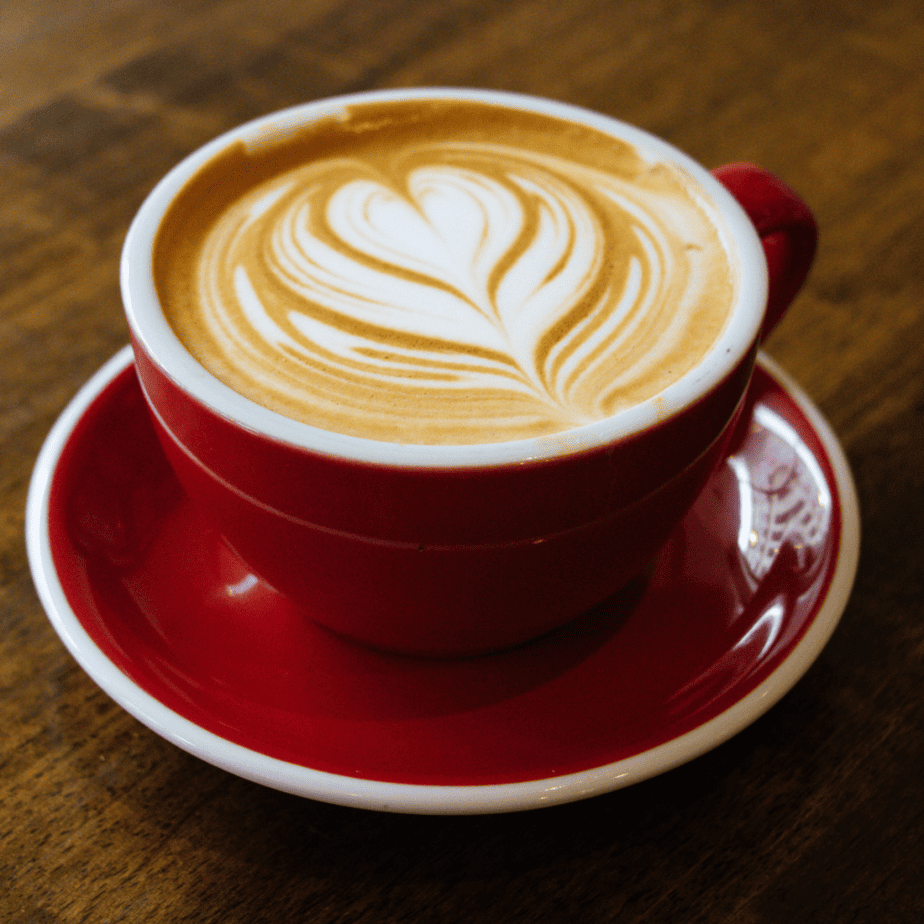 Cappuccino in a red cup that's on saucer on a wooden table.